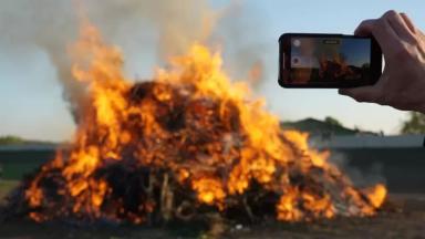 Gambar sebuah tumpukan benda yang terbakar dan tangan dari seseorang yang merekamnya menggunakan smartphone.