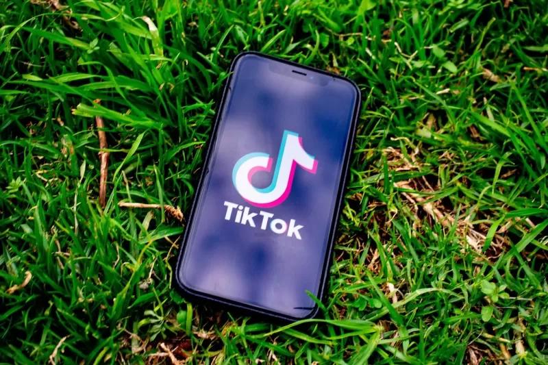 Gambar sebuah smartphone berisi logo TikTok di atas rumput berwarna hijau, ilustrasi TikTok Shop tidak tutup.