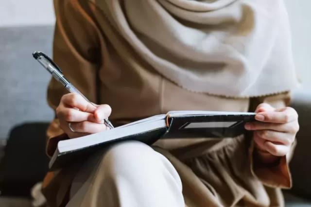 Gambar seorang wanita berhijab sedang menulis sesuatu di buku menggunakan pena.